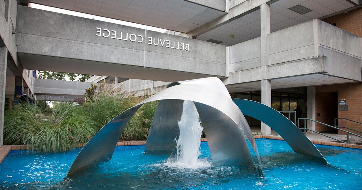 A large outdoor fountain with multiple silver arches and a single water spout in the center. The second level walkway is in the background with the Bellevue College sign in the center.